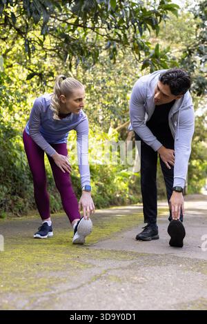 Amis divers effectuant des étirements de jambiers avant sur un sentier en forêt moussue avec des smartwatches Banque D'Images