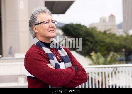 Homme senior debout avec les bras croisés sur la terrasse extérieure par balustrade, portant foulard et lunettes Banque D'Images