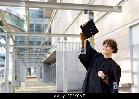 Blanc diplômé masculin debout sous la verrière dans la robe noire tenant chapeau au-dessus souriant, espace de copie Banque D'Images
