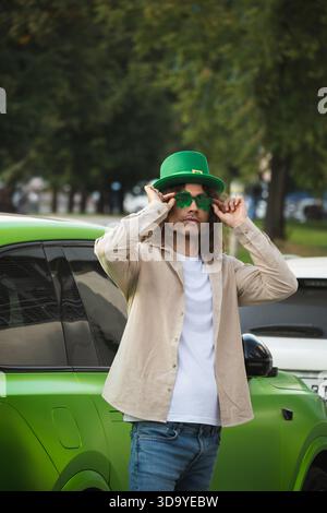 Élégant homme adulte caucasien des années 30 ajustant les lunettes de soleil de nouveauté verte et portant un chapeau de fête. Style de mode St patricks Day, portrait de style de vie urbain à Banque D'Images