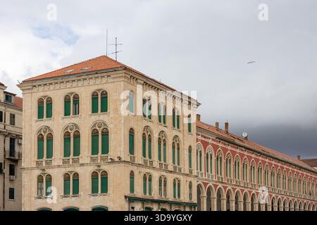 Un grand bâtiment historique ensoleillé avec des fenêtres cintrées, des volets verts et une longue arcade, transmettant le charme intemporel et l'architecture méditerranéenne Banque D'Images