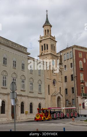 Une scène de rue européenne historique avec une église en pierre beige avec une grande tour de l'horloge, flanquée de vieux bâtiments. Un train touristique rouge vif se trouve dans Banque D'Images