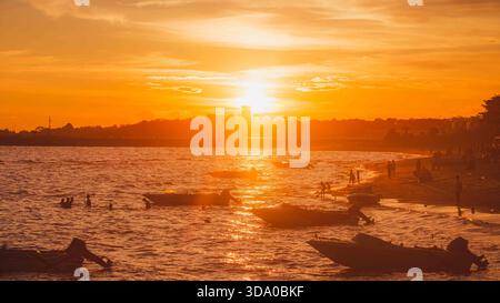 Un coucher de soleil orange et jaune éclatant sur le front de mer près de l'aéroport. De petits bateaux flottent dans l'eau chatoyante près d'une plage où les gens apprécient Banque D'Images