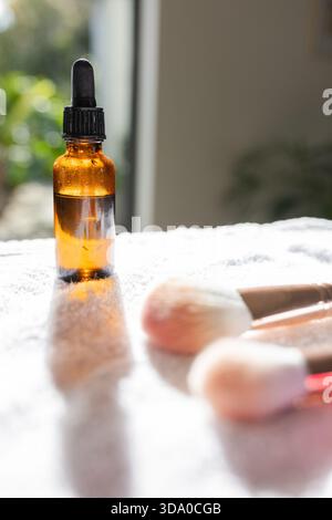 Bouteille compte-gouttes ambré reposant sur une serviette blanche avec condensation dans la lumière de fenêtre à côté des pinceaux de maquillage Banque D'Images