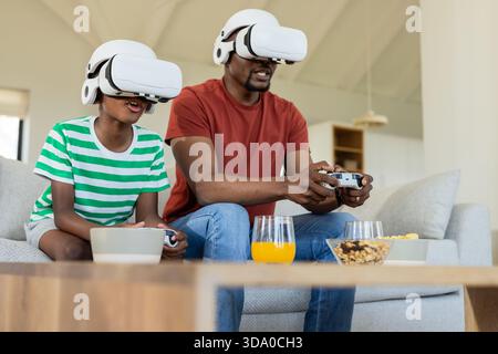 Père afro-américain et son fils portant des casques VR jouant à un jeu à la maison sur un canapé avec des contrôleurs Banque D'Images