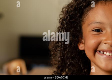 Fille afro-américaine souriante avec des cheveux bouclés près du moniteur de chaise en bois à la maison, espace de copie Banque D'Images