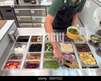 Une personne prépare une salade en utilisant une variété d'ingrédients frais, y compris des légumes verts, des tomates, des concombres et des pois chiches dans un environnement de cuisine. Banque D'Images
