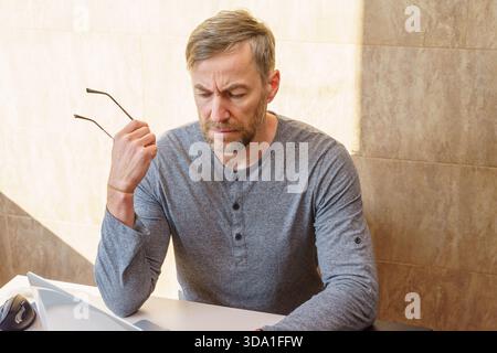 Un homme pensif d'âge moyen avec une barbe, portant une chemise grise, s'assoit à un bureau regardant vers le bas avec une expression concentrée tout en tenant ses lunettes. Banque D'Images