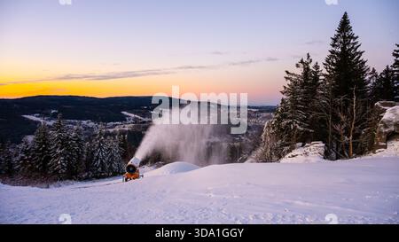 Un canon à neige fonctionne sur une pente enneigée, créant de la neige artificielle pour les skieurs. Le soleil se couche en arrière-plan, jetant de la lumière sur le paysage de montagne et les arbres. Banque D'Images