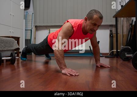 Homme athlétique d'âge moyen effectuant un exercice de push-up sur le sol de la salle de séjour avec un équipement de gymnastique à domicile Banque D'Images