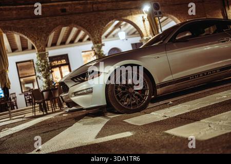 VO, italie 28 octobre 2025 : Mustang ford argenté garé dans une rue européenne historique la nuit, lampadaires soulignant les arches, les colonnes et le chro élégant Banque D'Images
