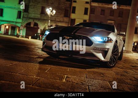VO, italie 28 octobre 2025 : ford Mustang avec des bandes de course stationnées sur la place pavée la nuit, illuminé les bâtiments historiques européens et les dramati Banque D'Images