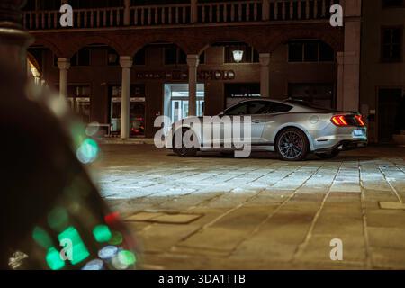 VO, italie 28 octobre 2025 : coupé ford Mustang argenté garé dans une rue illuminée de la ville la nuit, arches architecturales et lumières bokeh dans le backgroun Banque D'Images