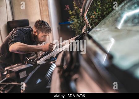 Rome, italie 20 novembre 2025 : mécanicien barbu en t-shirt inspectant et réparant un moteur de voiture à l'extérieur, axé sur la maintenance et l'automobile professionnelle Banque D'Images