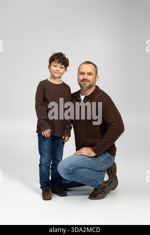 Un homme s'agenouille à côté de son fils souriant dans des chandails marron assortis dans un cadre intérieur confortable. Banque D'Images