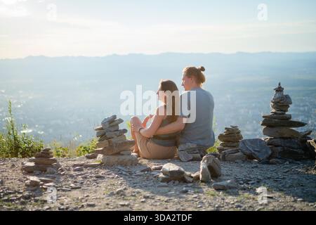 Couple romantique assis embrassé par derrière, regardant la vue imprenable sur la ville depuis le sommet de la montagne. Parfait pour les concepts de voyage, de randonnée et d'histoire d'amour. || autorisation du modèle Banque D'Images