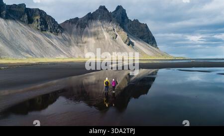 Deux aventuriers se tiennent sur la plage de sable noir sereine de Stokksnes, en Islande, contemplant la majestueuse montagne Vestrahorn et capturant les reflets dans des eaux calmes peu profondes au cours d'une journée paisible. || autorisation du modèle Banque D'Images
