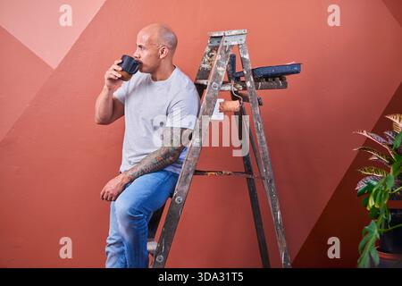 Homme avec des tatouages est assis sur une échelle patinée buvant du café près de la scène de rénovation Banque D'Images