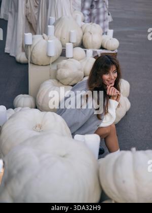 Belle jeune femme en pull blanc confortable et veste grise souriant et posant parmi de grandes citrouilles blanches et des bougies, capturant Hallowe joyeux Banque D'Images