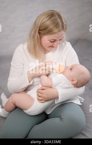 La femme est assise sur le lit, tient bébé dans ses bras et le nourrit du biberon. Concept de nourrir bébé avec du lait maternisé, alternative à l'allaitement maternel. Vertical Banque D'Images