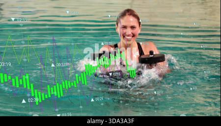 Femme souriante en maillot de bain noir soulevant des haltères en mousse et éclaboussant dans la piscine avec des graphiques financiers Banque D'Images