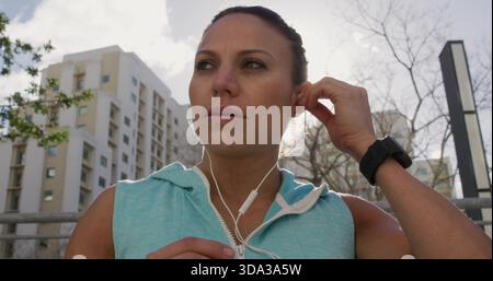 Femme ajustant tenant l'écouteur à l'oreille droite, câble de préhension, portant le dessus turquoise sur la rue urbaine Banque D'Images