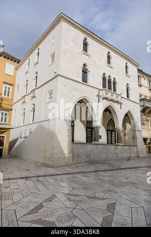 Un bâtiment historique en pierre blanche avec une élégante véranda voûtée se dresse sur une place de ville ensoleillée. Sa façade classique, ses détails en fer forgé et son pla carrelé Banque D'Images
