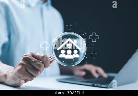 Concept d'assurance maladie familiale. Une personne tenant une loupe avec l'icône de la famille sous la maison symbolise l'assurance maladie familiale. croix médicale Banque D'Images