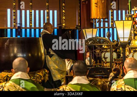 Service du matin à l'intérieur du hall principal (Hondo) du temple Zenkoji à Nagano : un prêtre dans un kesa noir se tient devant l'autel doré exécutant des actes rituels ; à droite, des objets rituels élaborés et utilisés pour orner l'autel peuvent être vus. La cérémonie quotidienne du matin à Zenkoji attire régulièrement des pèlerins de tout le Japon. Nagano, Préfecture de Nagano, Japon Banque D'Images