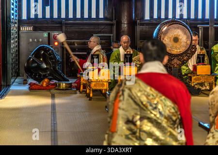Des moines dans le kesa cérémoniel effectuent la liturgie du matin dans le hall principal (Hondo) du temple Zenkoji à Nagano : un moine bat un grand tambour, tandis que d'autres s'assoient devant des lutrnes laqués noirs et dorés. Zenkoji abrite l’une des plus anciennes statues de Bouddha du Japon, qui, en tant que trésor national secret, est exposée au public tous les sept ans. Nagano, Préfecture de Nagano, Japon Banque D'Images