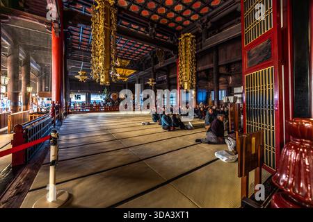 Une vue dans le hall principal (Hondo) du temple Zenkoji à Nagano lors de la cérémonie publique du matin. Les fidèles et les visiteurs vêtus de vêtements ordinaires et ordinaires sont assis sur le côté gauche du tatami ; plus loin dans le hall, des moines vêtus de robes d'or peuvent être vus devant l'autel. Les piliers laqués rouge foncé et or, le plafond à caissons peints et les rouleaux impériaux suspendus (makimono) reflètent le style de l'époque d'Edo, dont date l'Hondo actuel (1707). Nagano, Préfecture de Nagano, Japon Banque D'Images