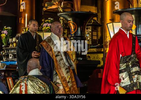 Cérémonie bouddhiste au temple Zenkoji à Nagano : un prêtre en chef, vêtu d'un magnifique kesa brodé d'or, tient une perle de prière juzu, flanquée de prêtres adjoints vêtus de noir et rouge. L'autel richement décoré en arrière-plan, avec des bougies, des offrandes florales et des objets rituels, est en harmonie avec le décor du Hondo, la salle principale de Zenkoji. Nagano, Préfecture de Nagano, Japon Banque D'Images