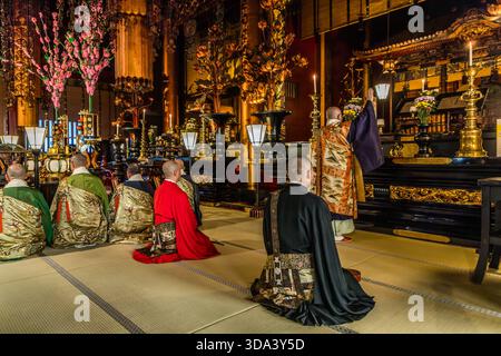 Plusieurs moines en kesa brodé d'or sont agenouillés sur des tatamis lors d'une cérémonie au temple Zenkoji à Nagano ; un prêtre exécute des actes rituels à l'autel doré, tandis que d'autres en robes rouges, vertes et dorées accompagnent la liturgie. Les fleurs de cerisier décoratives sur l'autel sont un élément décoratif typique des cérémonies Zenkoji. Nagano, Préfecture de Nagano, Japon Banque D'Images