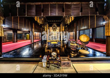Une vue symétrique dans le hall principal vide d'un temple bouddhiste à Nagano : l'allée centrale, en bois poli, mène à un autel doré ; à droite se dresse un grand gong circulaire (kin) sur un stand rouge. Des stores de paille (sudare) pendent du plafond ; sur le tapis de tatami au premier plan se trouvent un brûleur d'encens, des boîtes laquées et des offrandes. Les zones recouvertes de moquette rouge à gauche et à droite marquent les places assises pour les fidèles. Nagano, Préfecture de Nagano, Japon Banque D'Images