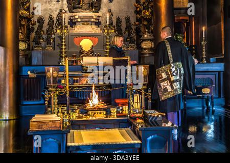Un visiteur occidental se tient devant un autel de flamme dans une chapelle d'un sous-temple Zenkoji à Nagano ; un moine à droite explique le rituel et sa signification. Le feu sur l'autel (feu rituel de goma ou cérémonie d'allumage des bougies) est une caractéristique des cérémonies quotidiennes dans les temples bouddhistes. Nagano, Préfecture de Nagano, Japon Banque D'Images