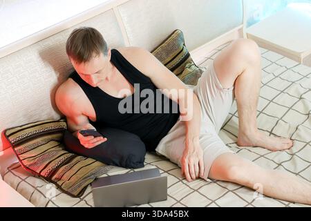 Dans une chambre tranquille, un homme en débardeur et short se repose sur des oreillers, engagé avec son téléphone tandis qu'un ordinateur portable est assis fermé sur le lit dans une ambiance douce Banque D'Images