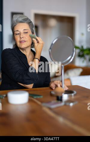 Femme mature pratiquant le massage facial avec une pierre gua Sha, favorisant le rajeunissement de la peau et la relaxation dans un cadre familial Banque D'Images