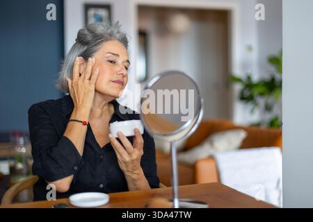 Femme mature prenant soin de sa peau, appliquant une crème anti-âge tout en regardant dans un petit miroir Banque D'Images