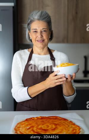 Femme âgée portant un tablier, tenant un bol de fromage râpé, debout dans une cuisine moderne tout en faisant de la pizza Banque D'Images