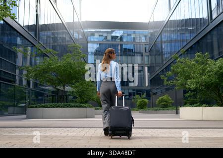 Vue arrière d'une femme confiante tirant une valise à travers un quartier d'affaires moderne. Bâtiments en verre réfléchissant, cadre urbain, concept de voyage, CA Banque D'Images