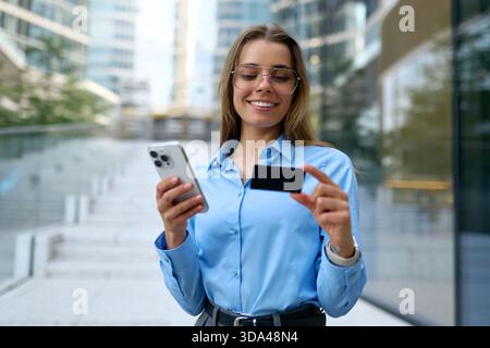 Jeune femme d'affaires en chemise bleue tenant smartphone et carte de crédit, souriant tout en effectuant un paiement en ligne à l'extérieur dans un cadre urbain moderne. Conce Banque D'Images