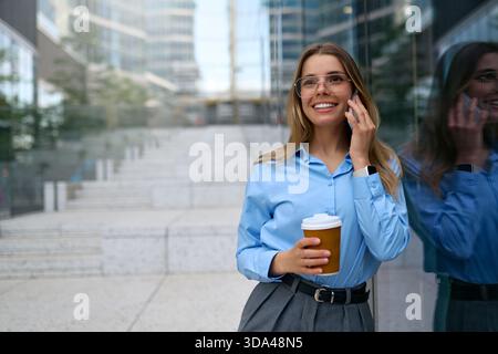 Jeune femme d'affaires confiante en chemise bleue et lunettes tenant le café, parlant sur smartphone, debout à côté d'un mur de verre avec réflexion en urbain Banque D'Images