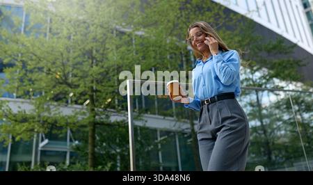 Jeune femme souriante en chemise bleue, tenant une tasse à café et parlant au téléphone tout en descendant des escaliers en marbre dans un quartier d'affaires moderne. La vie urbaine Banque D'Images