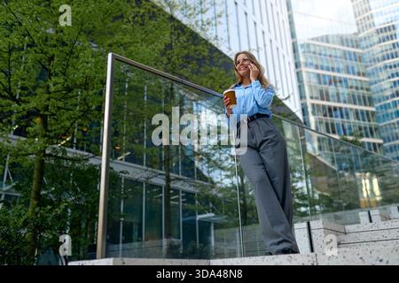 Jeune femme confiante en chemise bleue et lunettes appuyée sur la balustrade en verre, tenant le café et parlant sur smartphone à l'extérieur dans les affaires urbaines modernes d Banque D'Images