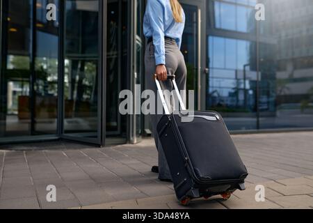 Vue arrière d'une femme en tenue formelle tirant la valise noire vers l'entrée de l'immeuble de bureaux en verre. Voyage d'affaires, voyage d'affaires, vie urbaine moderne Banque D'Images