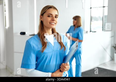 Dans un environnement de soins lumineux, une infirmière confiante en bleu frotte sourit tout en tenant une planche à pince, et un autre membre du personnel travaille pour s'assurer que les patients f Banque D'Images
