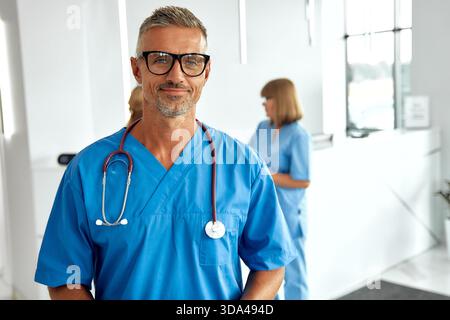 Un médecin masculin très confiant, vêtu de gommages bleus et portant des lunettes, se tient dans un environnement de soins de santé contemporain, entièrement prêt à aider Banque D'Images
