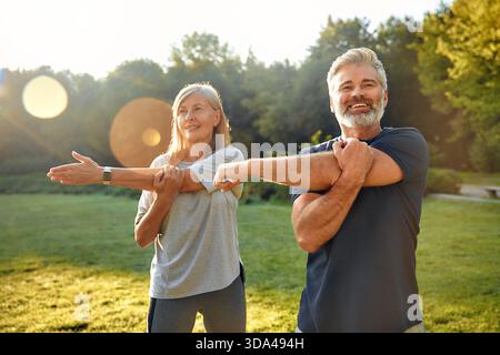Un couple joyeux s'étire ensemble à l'extérieur dans un parc serein alors que le soleil se couche, favorisant la forme physique et le bien-être tout en profitant pleinement de la compagnie de l'autre Banque D'Images
