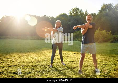 Un couple joyeux s'étire ensemble à l'extérieur dans un parc serein alors que le soleil se couche, favorisant la forme physique et le bien-être tout en profitant pleinement de la compagnie de l'autre Banque D'Images