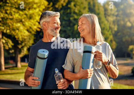 Un couple mature en forme et heureux profite d'une belle journée ensoleillée en plein air dans un joli parc, tenant joyeusement leurs tapis de yoga et bouteilles d'eau pendant eng Banque D'Images